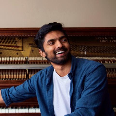 Kalaisan sits in front of an upright piano. The front is taken off so we can see the inside string and mallets. He is sm