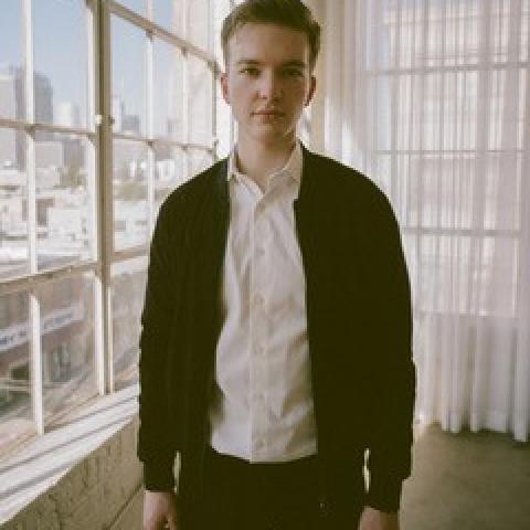 Misha stands in front of a window. In the background is a city scape. He wears a white shirt and black jacket. 