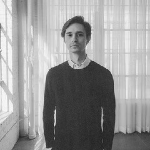 A black and white photo of Nathan standing in front of a window with a white sheer curtain. Sun beams can be seen coming