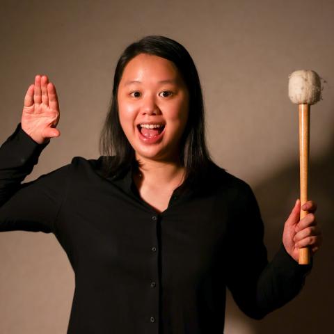 Hoi Tong Keung stands with a drum mallet and left hand out stretched. She smiles in front of a grey background. 