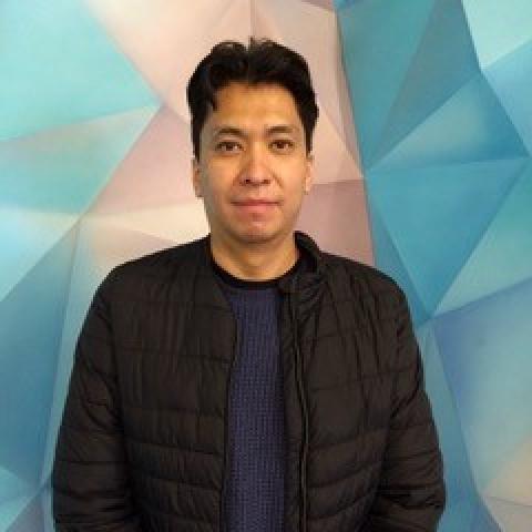 Diego stands in front of a blue and pink geometric wall. He is wearing a black jacket. 