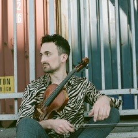 Michael sits in a patterned shirt in front of shipping containers holding a violin. 
