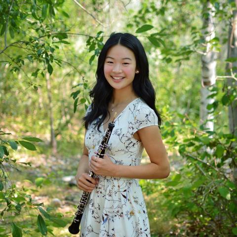 Woman standing on forest path in a floral dress holding an oboe in front of her