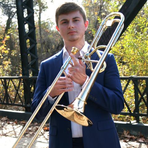 Man in blue jacket holding a trombone as if about to play while standing on a metal frame bridge with trees in the backg