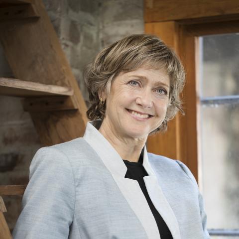 Woman leaning against a wooden ladder smiling broadly at the camera in a blazer with popped collar
