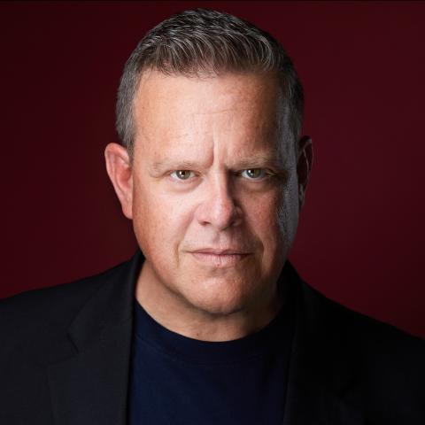 Man in dark clothing with short cropped blonde hair in front of burgundy backdrop 