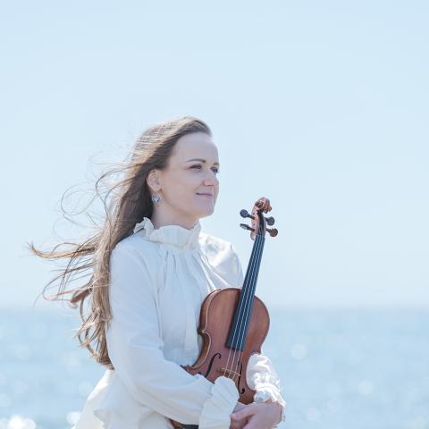 Woman standing in front of water in flowy clothes holding violin