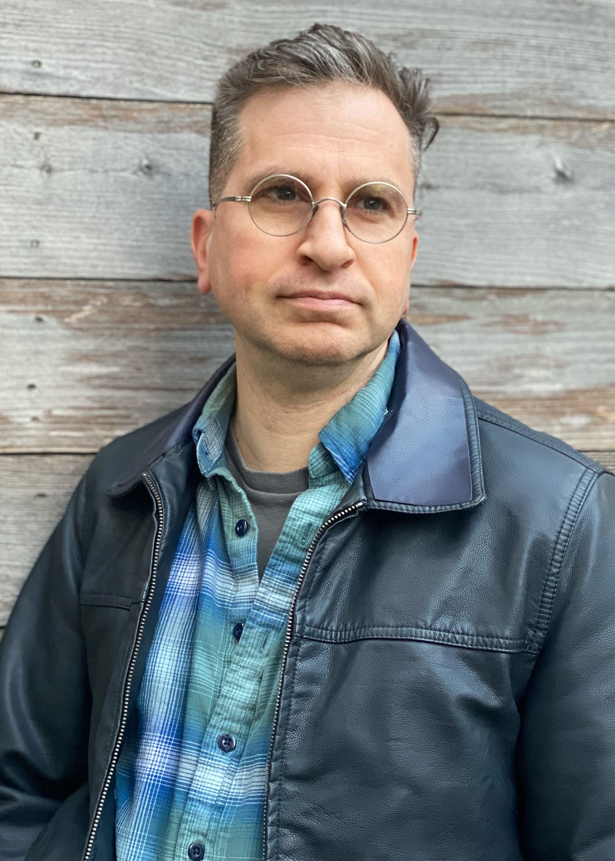 Man standing against wooden wall wearing a plaid shirt and leather jacket